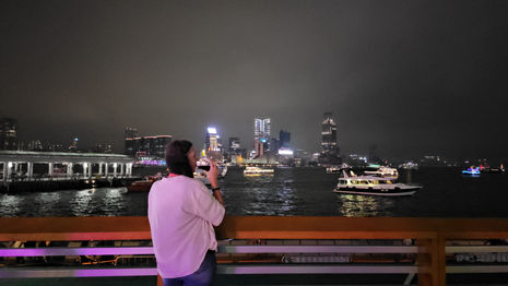 A person in a white shirt stands on a waterfront, taking a photo of a brightly lit city skyline at night. Boats with lights are visible on the water.