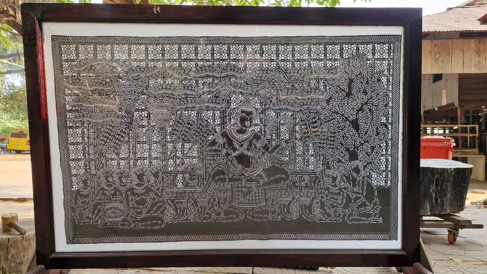 A framed, intricate carving on a metallic sheet depicts a detailed scene with various figures and patterns. The carving is displayed outdoors, with trees and a building visible in the background.