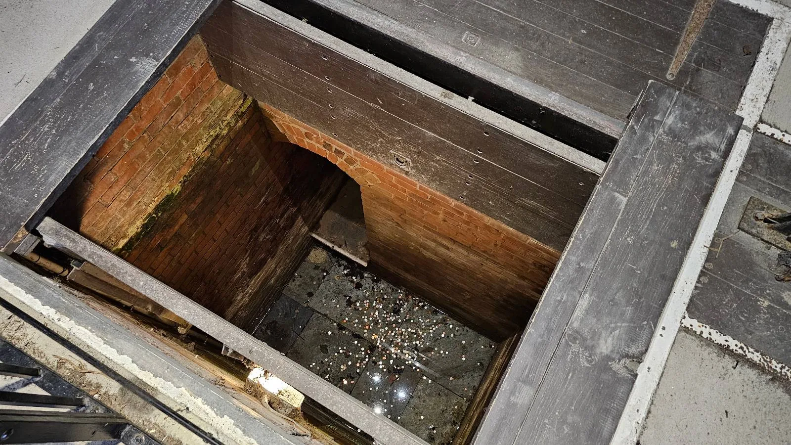 A view looking down into a deep, narrow pit lined with wooden panels and filled with coins at the bottom.