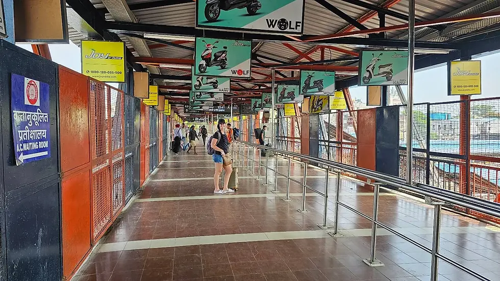 Train station walkway with a woman standing, pulling a suitcase. Advertisements for scooters hang above. Signs point to an A.C. waiting room.