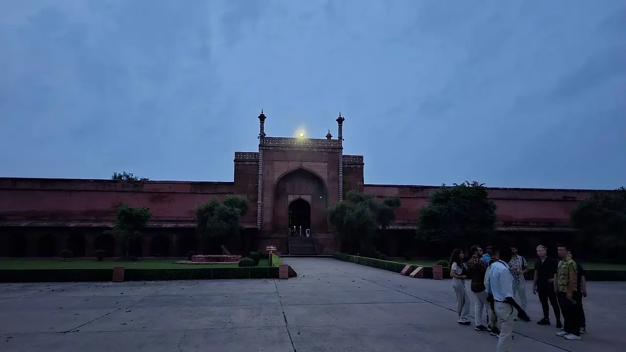 A group of people stands near the entrance of a large red-brick building with an arched gateway, illuminated by a light at dusk, with a cloudy sky in the background.