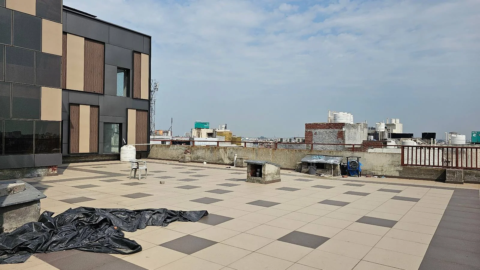 A spacious rooftop terrace with beige and brown tiles, a few scattered chairs and tables, and some equipment. Part of a modern building is on the left, with a cityscape and cloudy sky in the background.