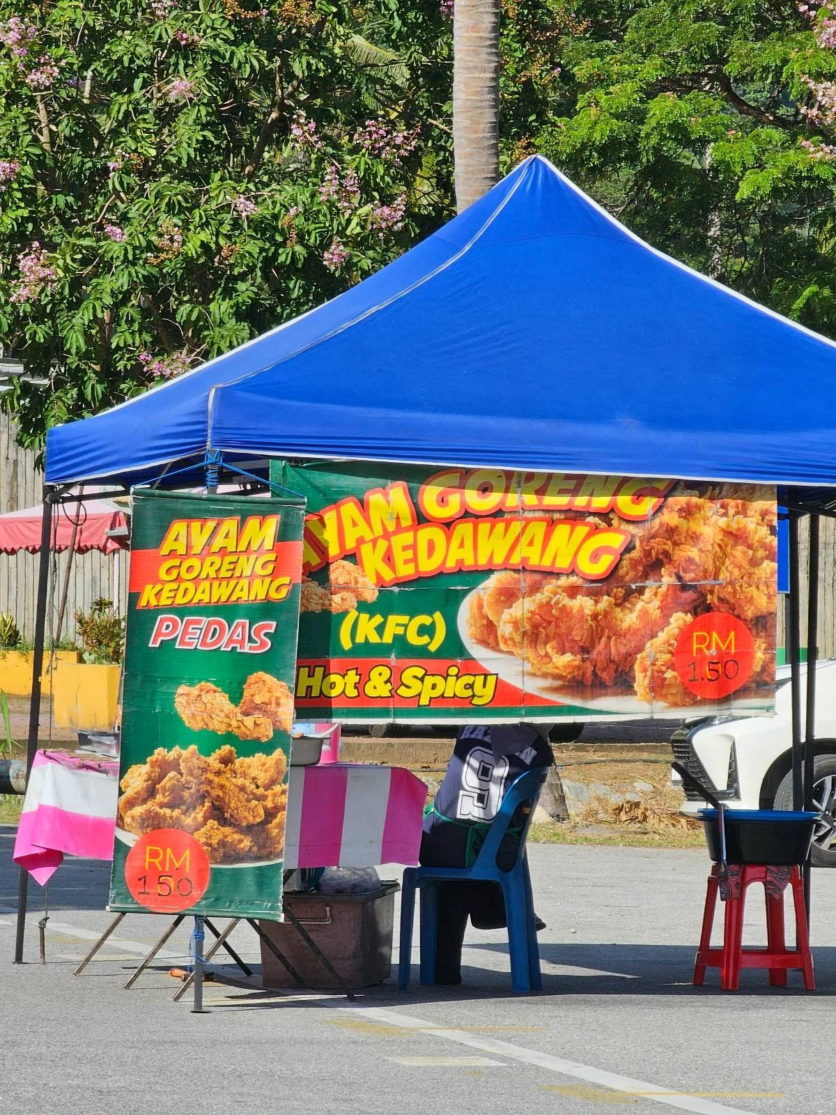A blue canopy shading a street food stall with a colorful banner advertising "Ayam Goreng Kedawang" (KFC) as hot and spicy. The setup includes a table with a pink and white striped cloth and chairs on a sunny day.