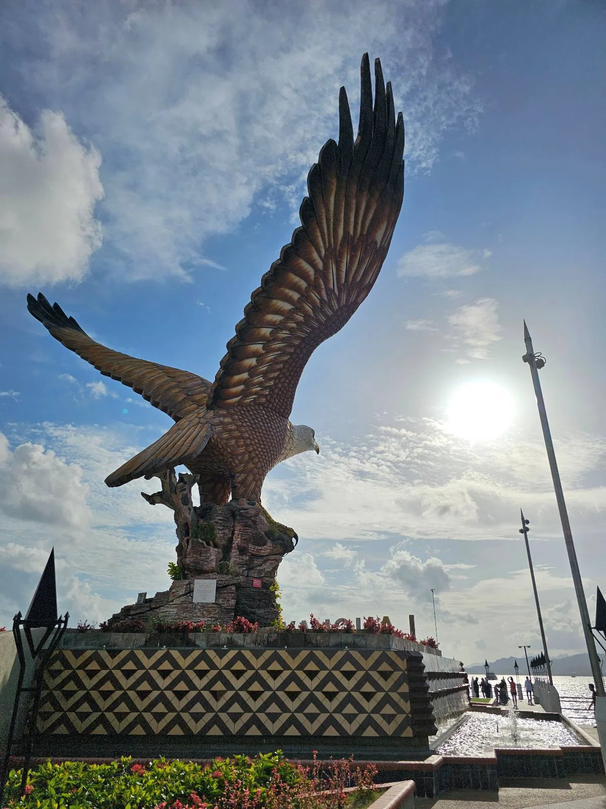 A large eagle statue with outstretched wings stands on a decorated pedestal under a bright, partly cloudy sky. People are gathered around the base, and the sun shines in the background.