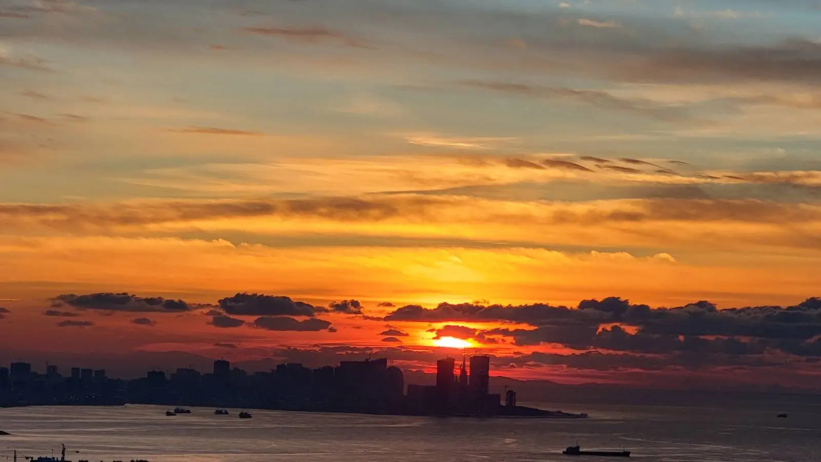 Sunset over a city skyline with buildings silhouetted against an orange and blue sky, clouds scattered above, and a calm body of water in the foreground.
