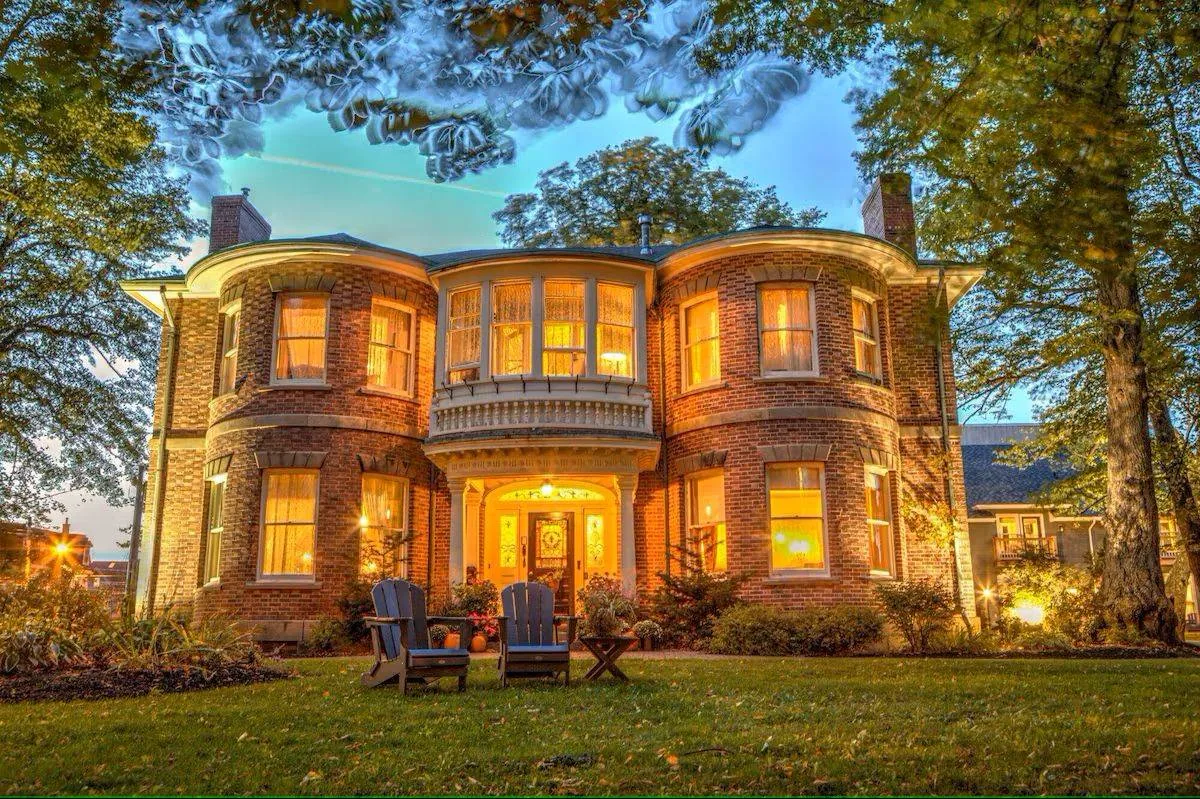 A large, illuminated two-story brick house with many windows, surrounded by trees and situated on a well-manicured lawn. Two wooden chairs are placed on the grass in the front yard, under the twilight sky.