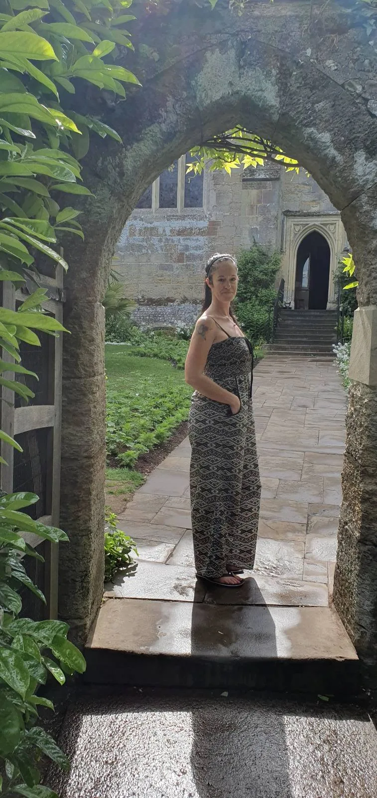 A person in a long dress stands under a stone archway, surrounded by lush greenery. In the background, there's a stone building with an open doorway. The ground is wet, suggesting recent rain.