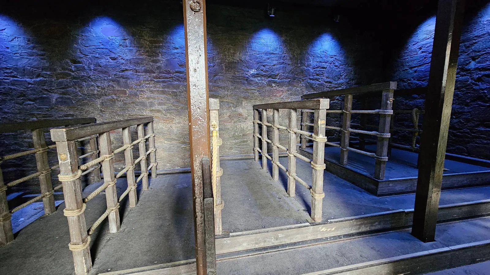A dimly lit room with stone walls and three wooden-railed platforms, separated by metal bars, illuminated by blue overhead lights, creating a somber and industrial atmosphere.