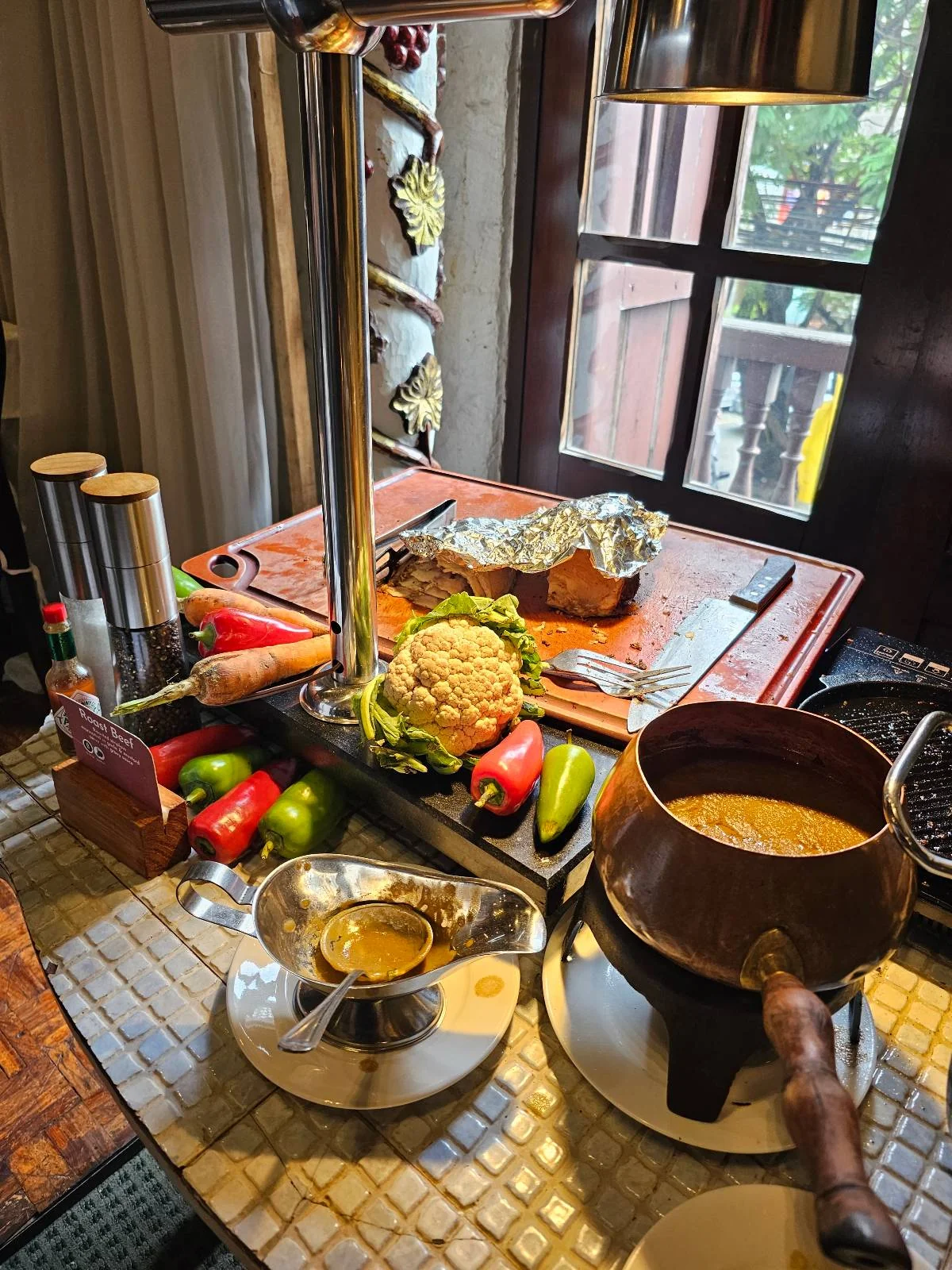 Table with fondue pot, cheese sauce, bread, vegetables, and cutting board with foil-wrapped food. Nearby are condiments and various peppers, with a window in the background.