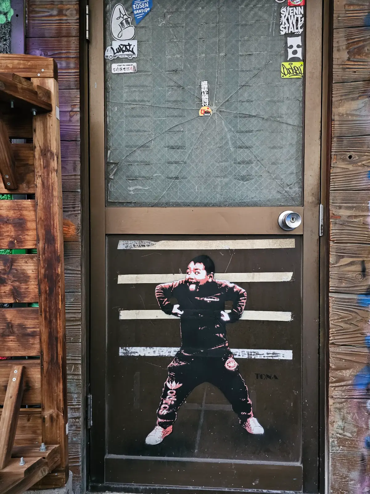 A metal door with a mesh window is covered in stickers and features a large street art image of a person in black clothing, with arms raised and a surprised expression, pasted across the lower panels.