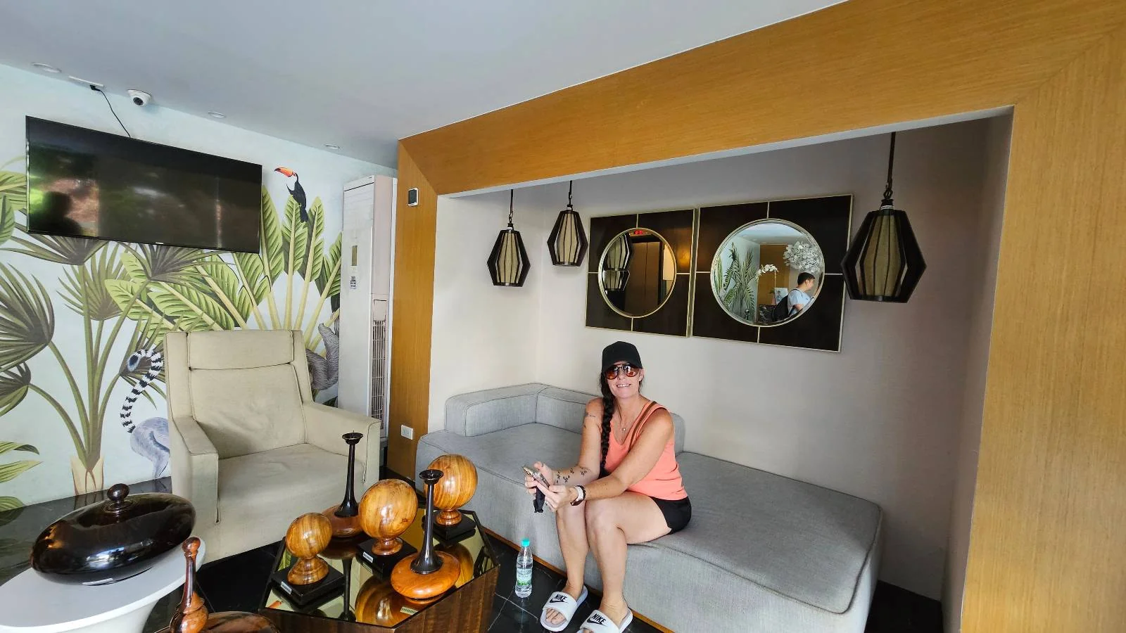 A woman sits on a gray couch in a modern living room with decorative mirrors, hanging lights, tropical wallpaper, and a coffee table with wooden ornaments.