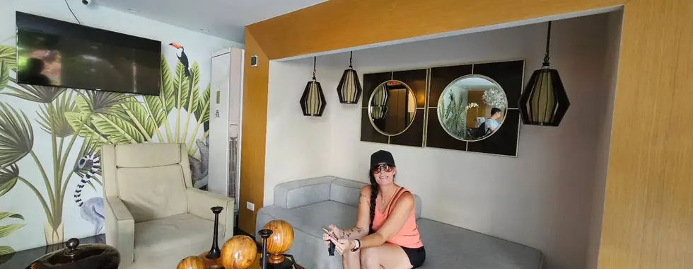 A woman sits on a gray couch in a modern living room with decorative mirrors, hanging lights, tropical wallpaper, and a coffee table with wooden ornaments.