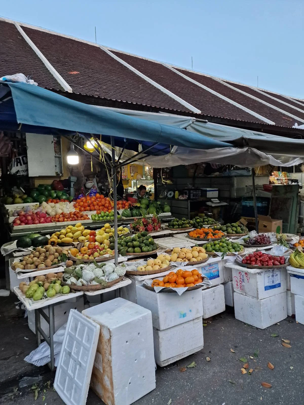 A bustling outdoor market stall displays a colorful variety of fruits and vegetables, including oranges, bananas, and tomatoes, arranged in baskets and crates under a canopy with a corrugated roof.