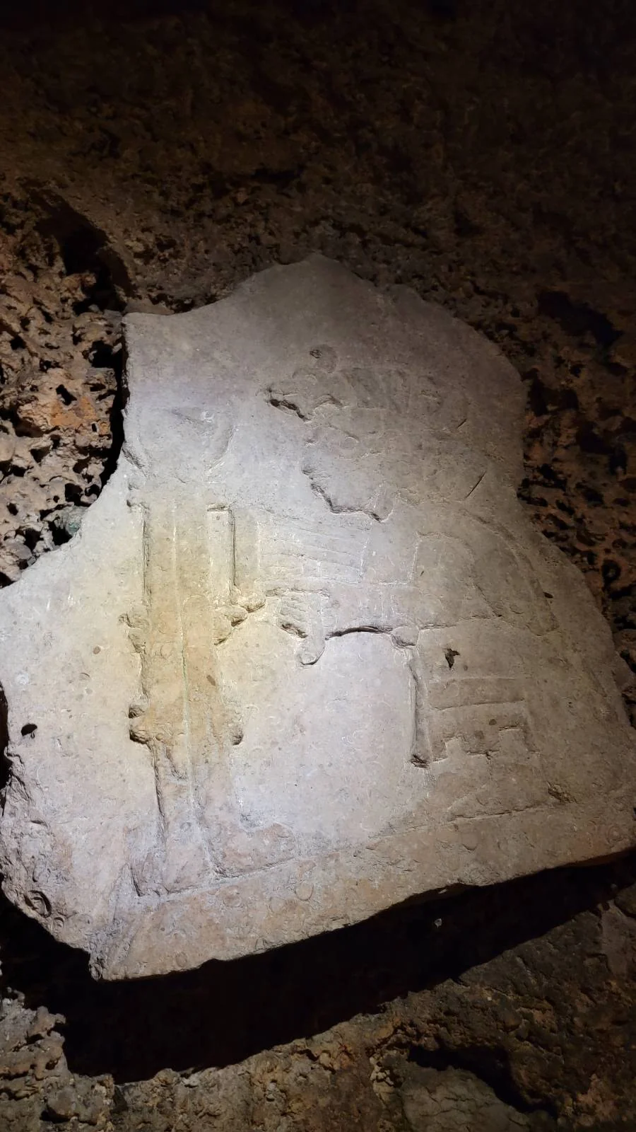 A piece of weathered, carved limestone with faint ancient engravings, lying on a rough, textured surface. Shadows and lighting create contrasts, highlighting the worn details and irregular shape of the stone.