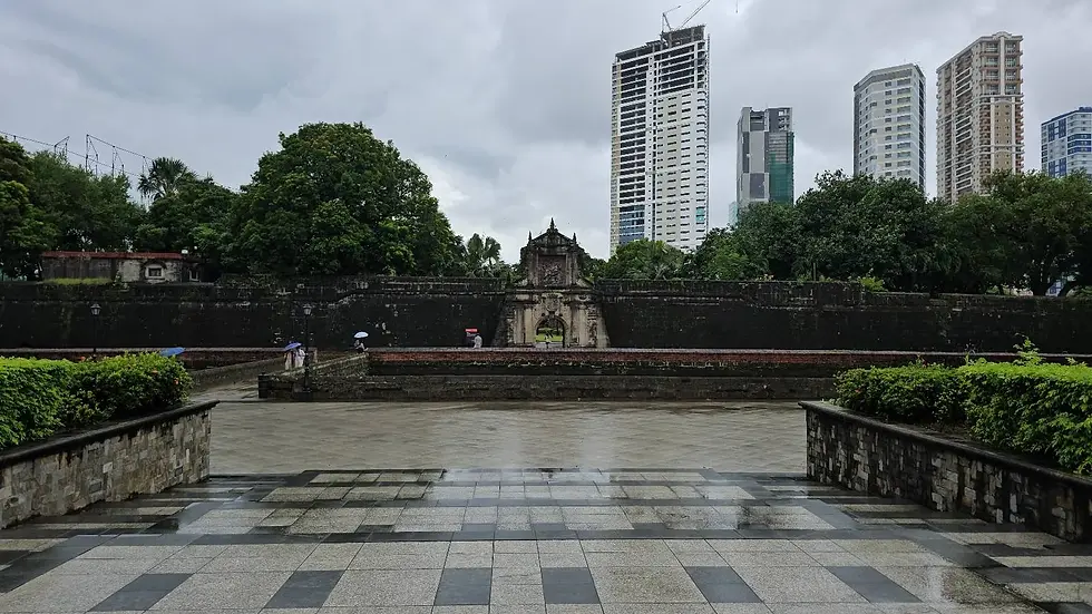 Historic stone gate with people under umbrellas. Overcast sky, lush green trees, and modern skyscrapers in the background. Wet pathway.