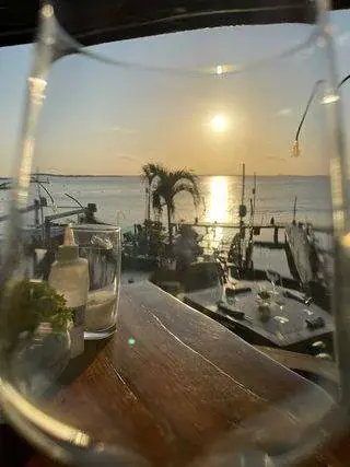 A scenic view of a sunset over a body of water, seen through an empty wine glass. The sunlight reflects on the water, and palm trees are visible near the shoreline. Tables with dining settings are in the foreground.