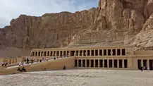 Tourists visiting the ancient Egyptian temple of Hatshepsut, a large, terraced structure embedded in a cliff face under a clear sky.