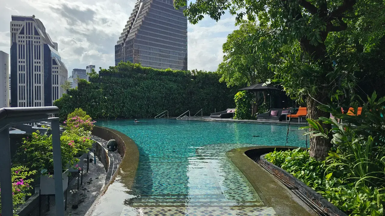 Curved infinity pool surrounded by trees and Bangkok high-rise buildings.