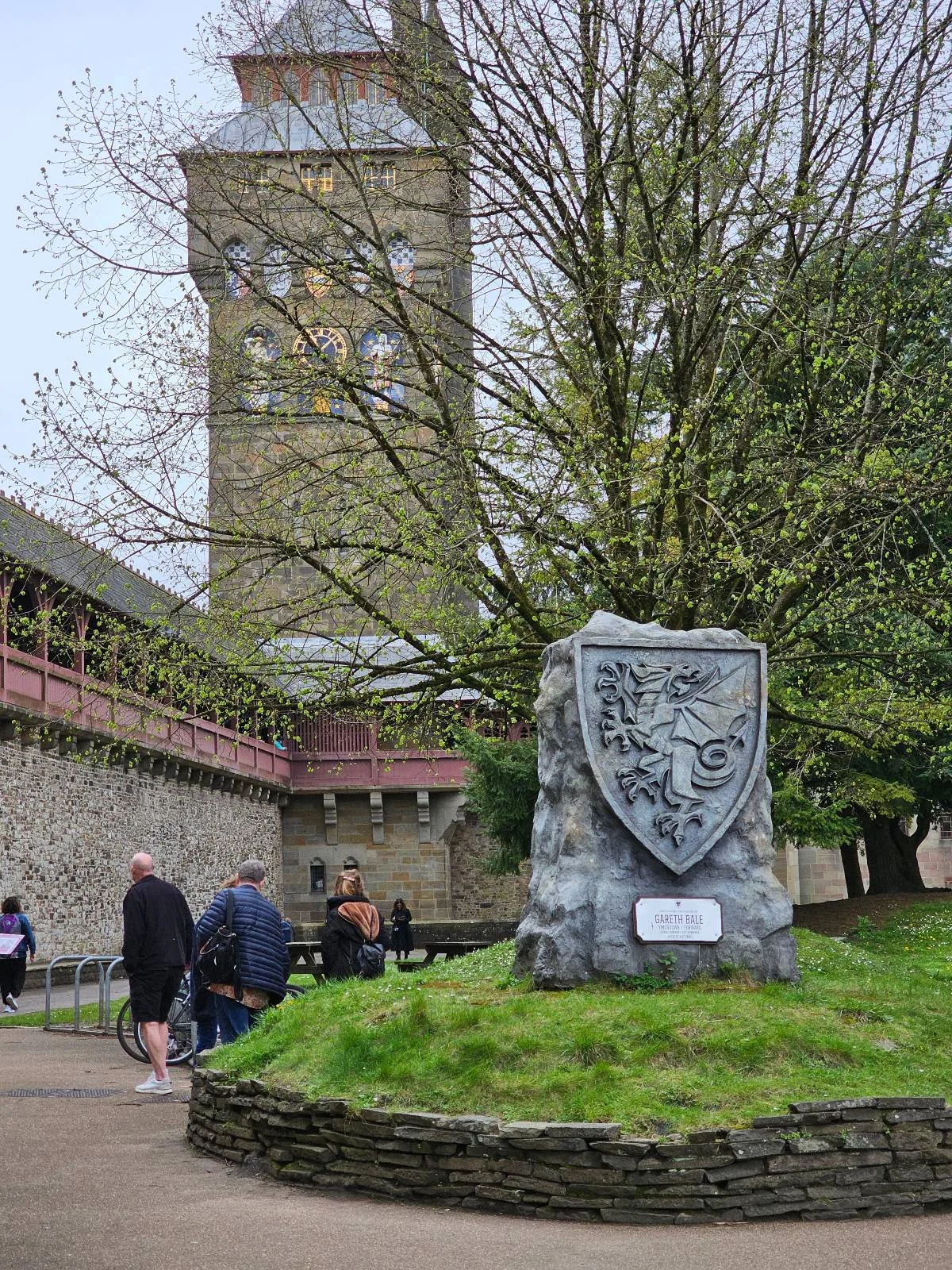A stone monument featuring a large shield with a coat of arms in front of a church tower, surrounded by lush greenery and several people walking by.