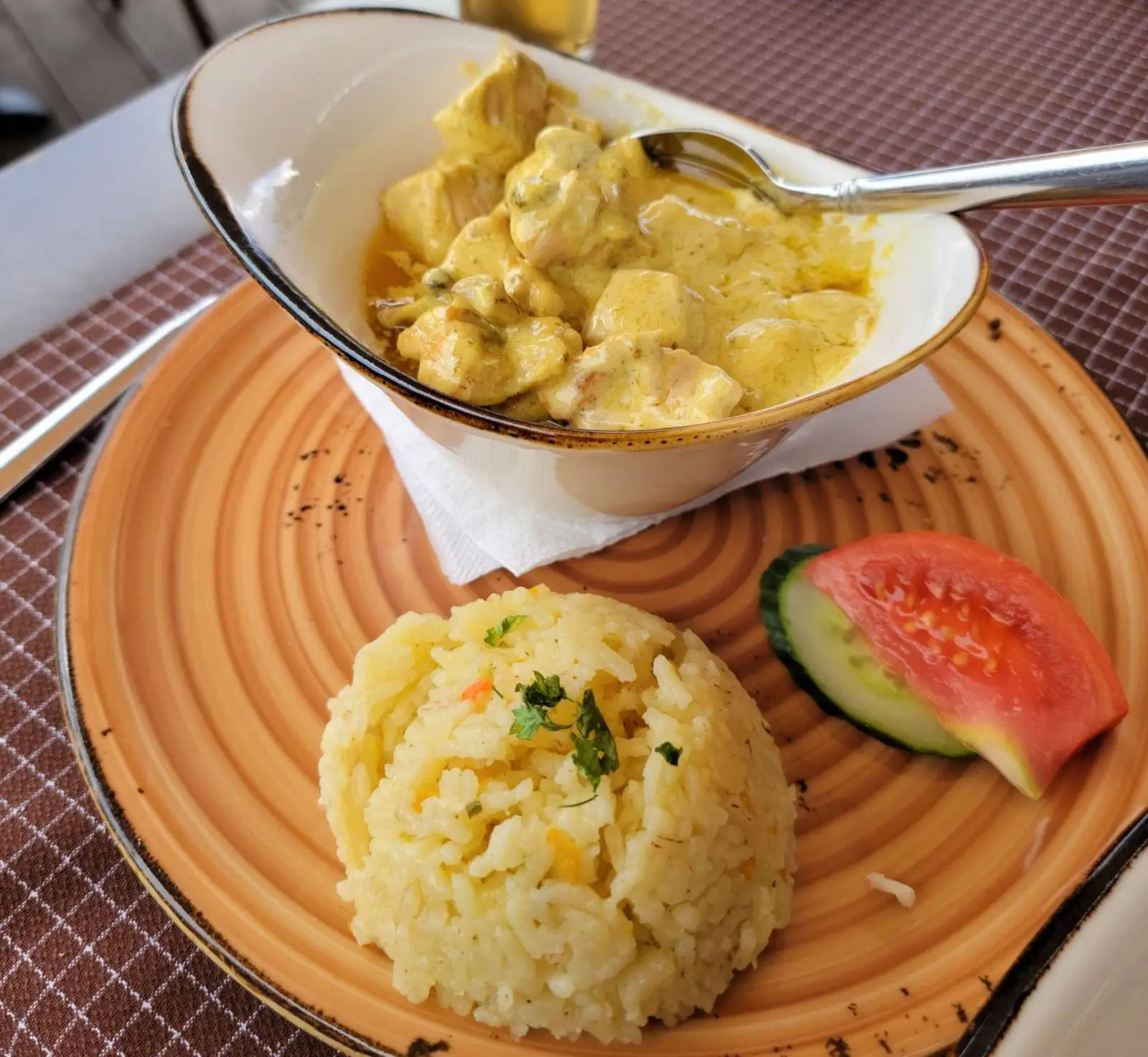 A plate with a serving of yellow rice garnished with parsley, a slice of cucumber, and a wedge of tomato. Beside it, there's a bowl of chicken curry with a creamy yellow sauce and a spoon resting on the side.