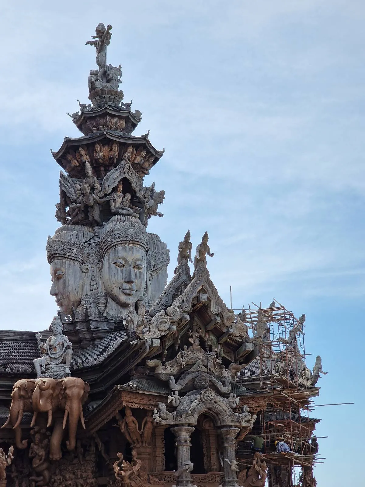 Intricate wooden carvings and sculptures adorn the roof of a traditional temple, featuring multiple detailed figures and ornate designs against a clear blue sky. Scaffolding is visible on the right side of the structure.