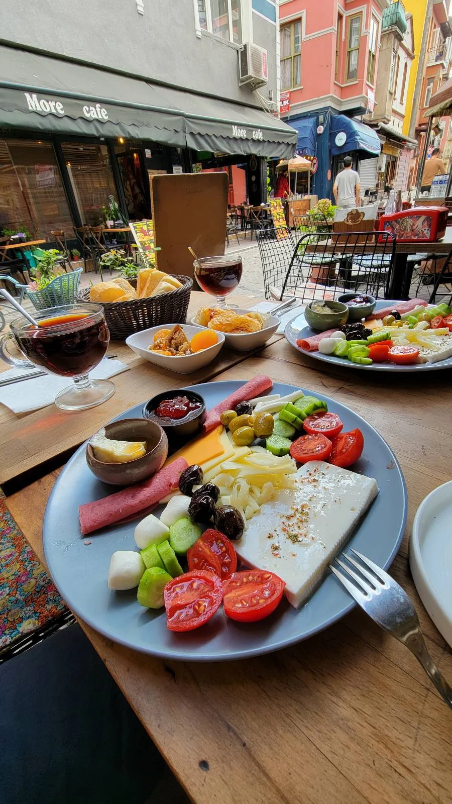 A platter with cheese, salami, cucumbers, olives, tomatoes, and jam. A bowl of bread and a drink are in the background.