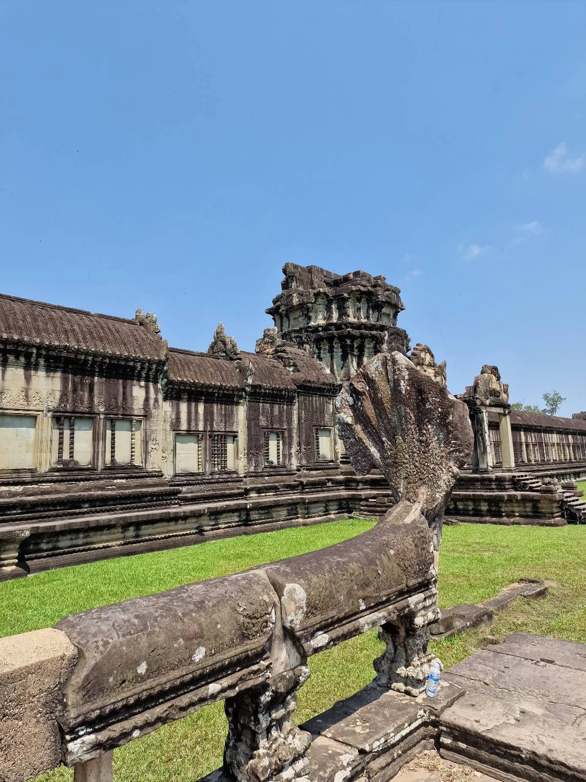 Ancient stone ruins of Angkor Wat temple under a clear blue sky, with a weathered, serpent-shaped balustrade in the foreground and grassy courtyard visible.