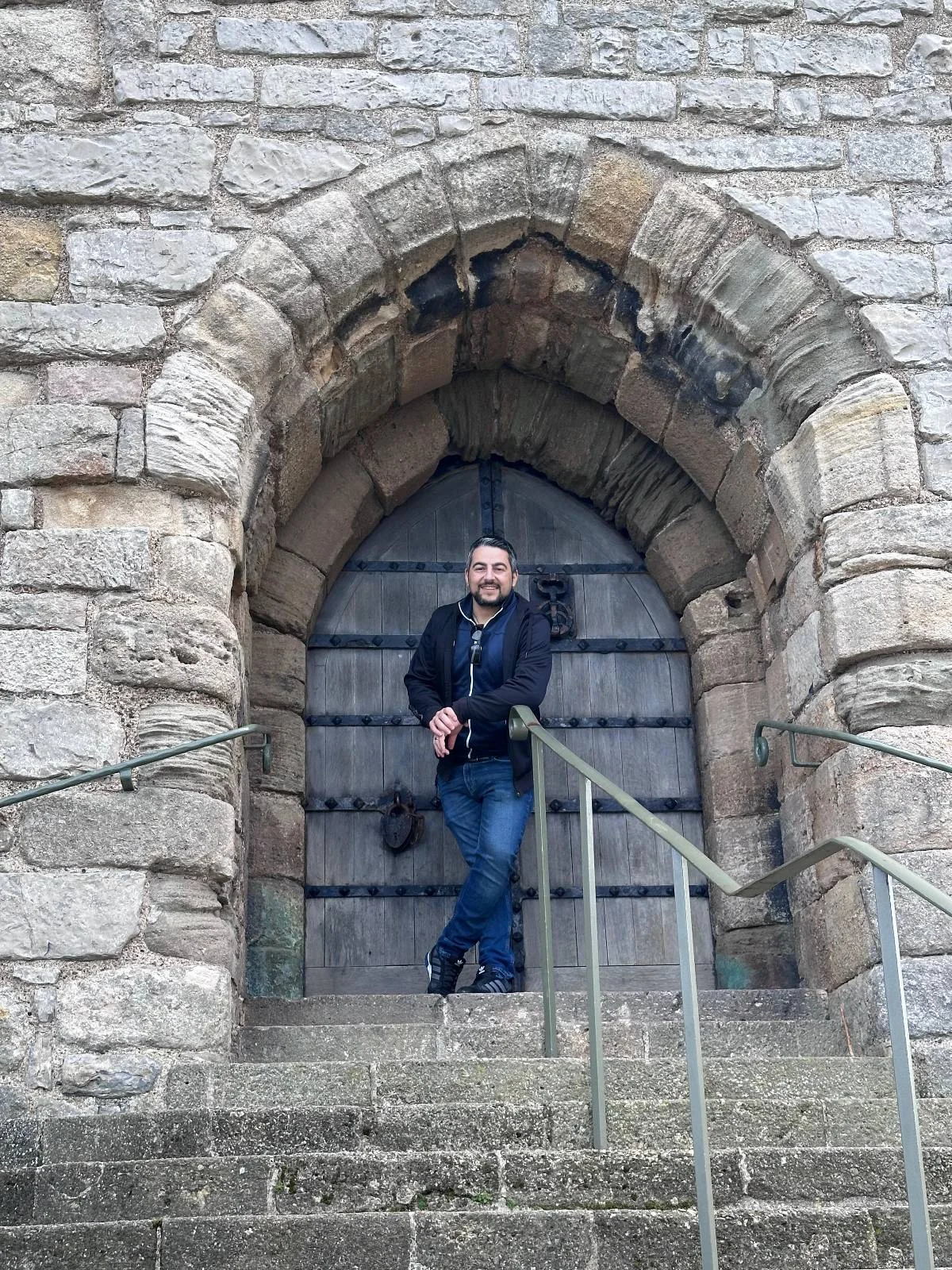 A person stands on stone steps in front of a large, arched wooden door set in a stone wall, holding a bag and looking at the camera. Metal railings line the steps.
