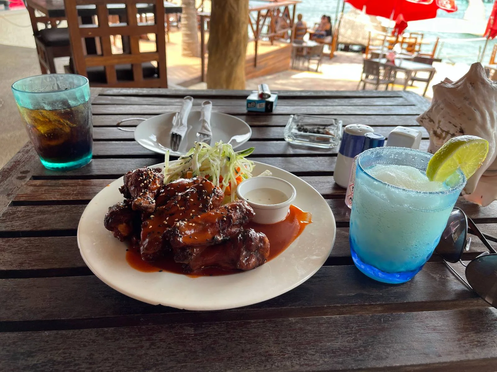 A plate of sauced chicken wings with a side of salad and dip sits on a wooden table. Two drinks, a clear soda with ice and a blue-rimmed glass with a lime wedge, are nearby. Outdoor seating and beach are in the background.