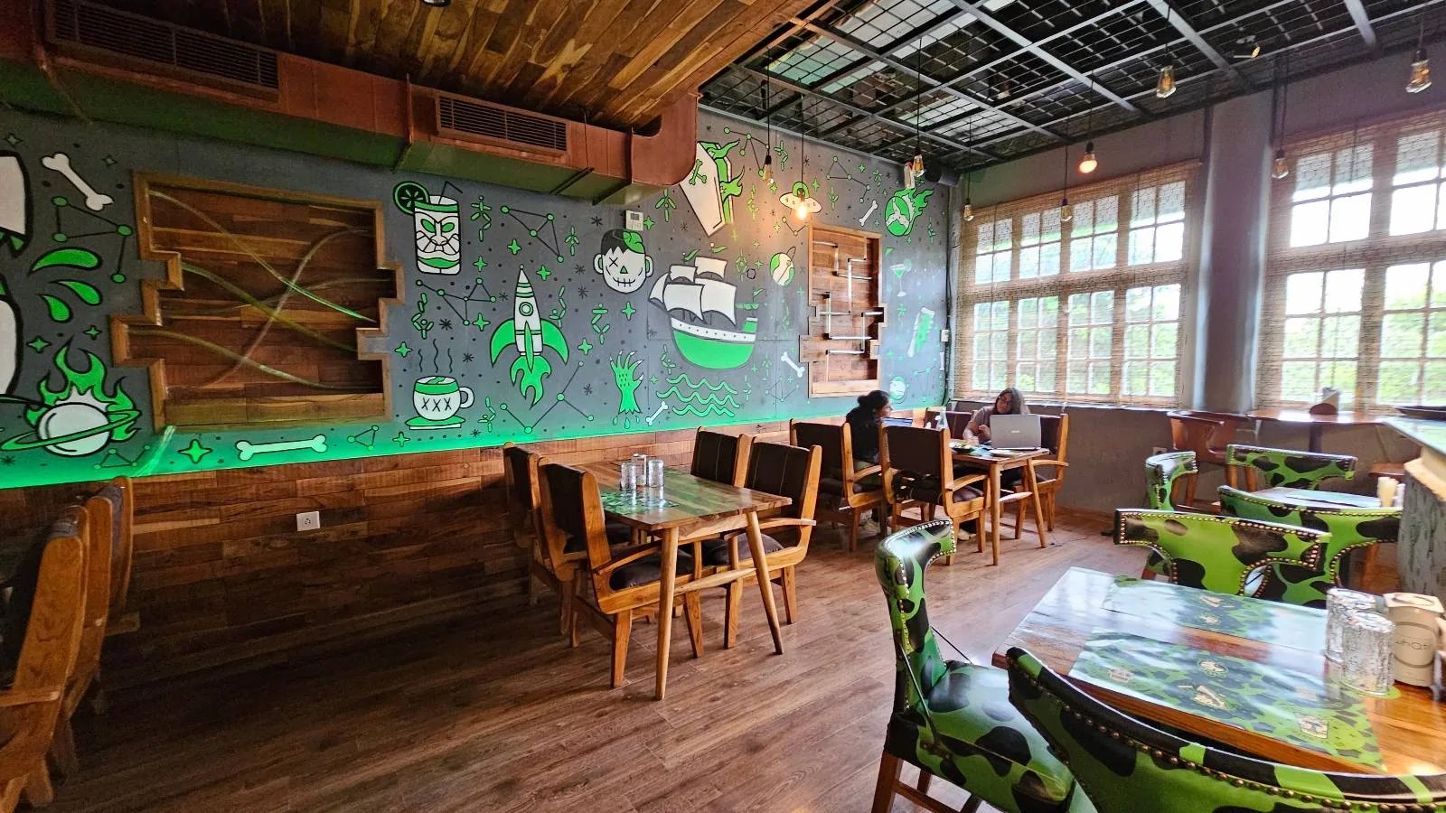 A cozy cafe with wooden furniture, green and pirate-themed decor. Walls feature nautical designs and a ship mural. Large windows let in natural light. Tables and chairs are arranged around the room, with plants adding a fresh touch.
