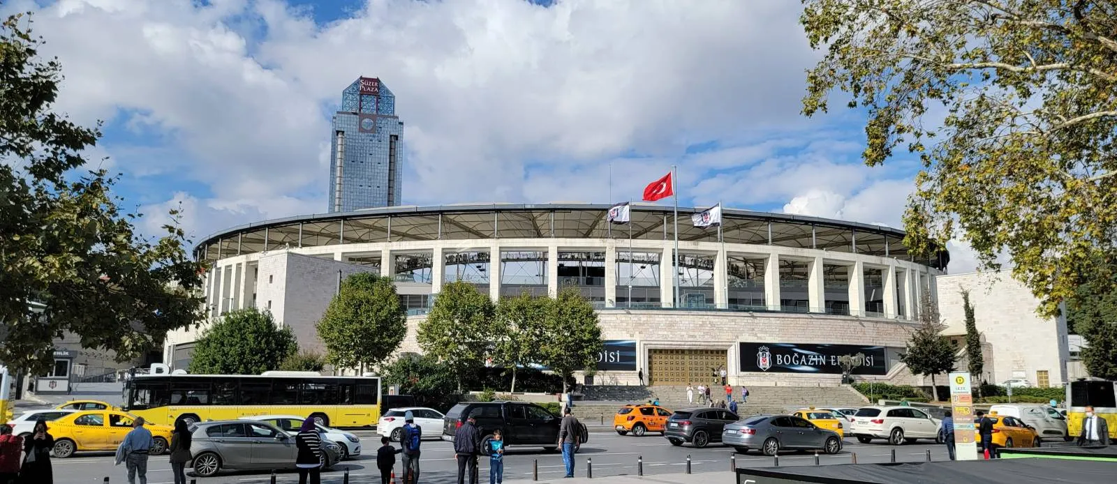 A stadium with a Turkish flag on top, surrounded by trees and parked cars, including yellow taxis and buses. People walk along the sidewalk under a partly cloudy sky. A tall modern building rises in the background.