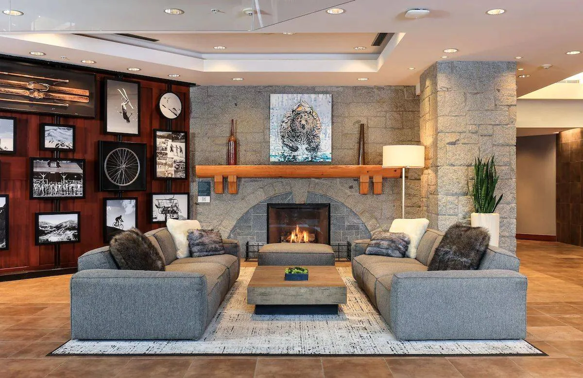 A cozy reception area with a modern fireplace at the center, surrounded by stone walls. Two gray sofas face each other over a rectangular coffee table. The walls are decorated with framed photos and art. A large plant stands near a floor lamp on the right.