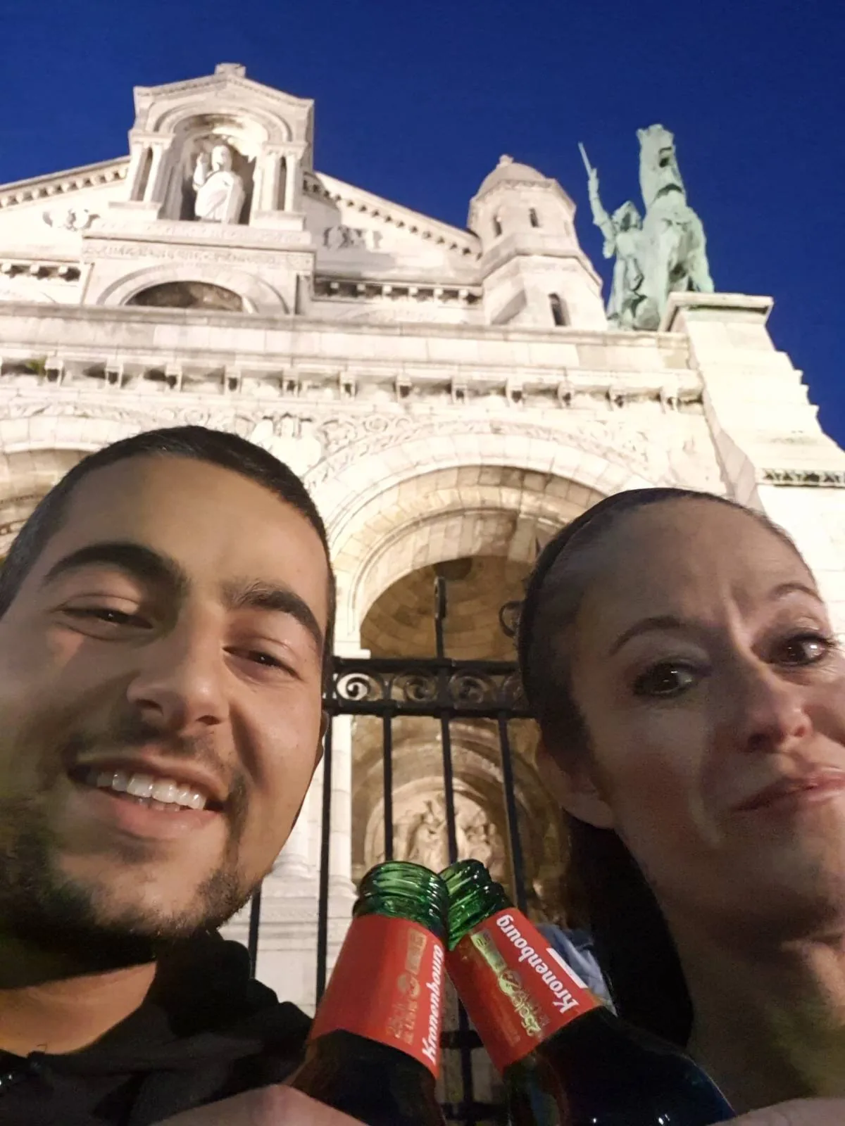 A man and woman smile while holding up two bottles in front of an ornate stone building with statues. The sky is clear and deep blue, suggesting it's evening or nighttime. The angle is from below, making the building appear grand and imposing.