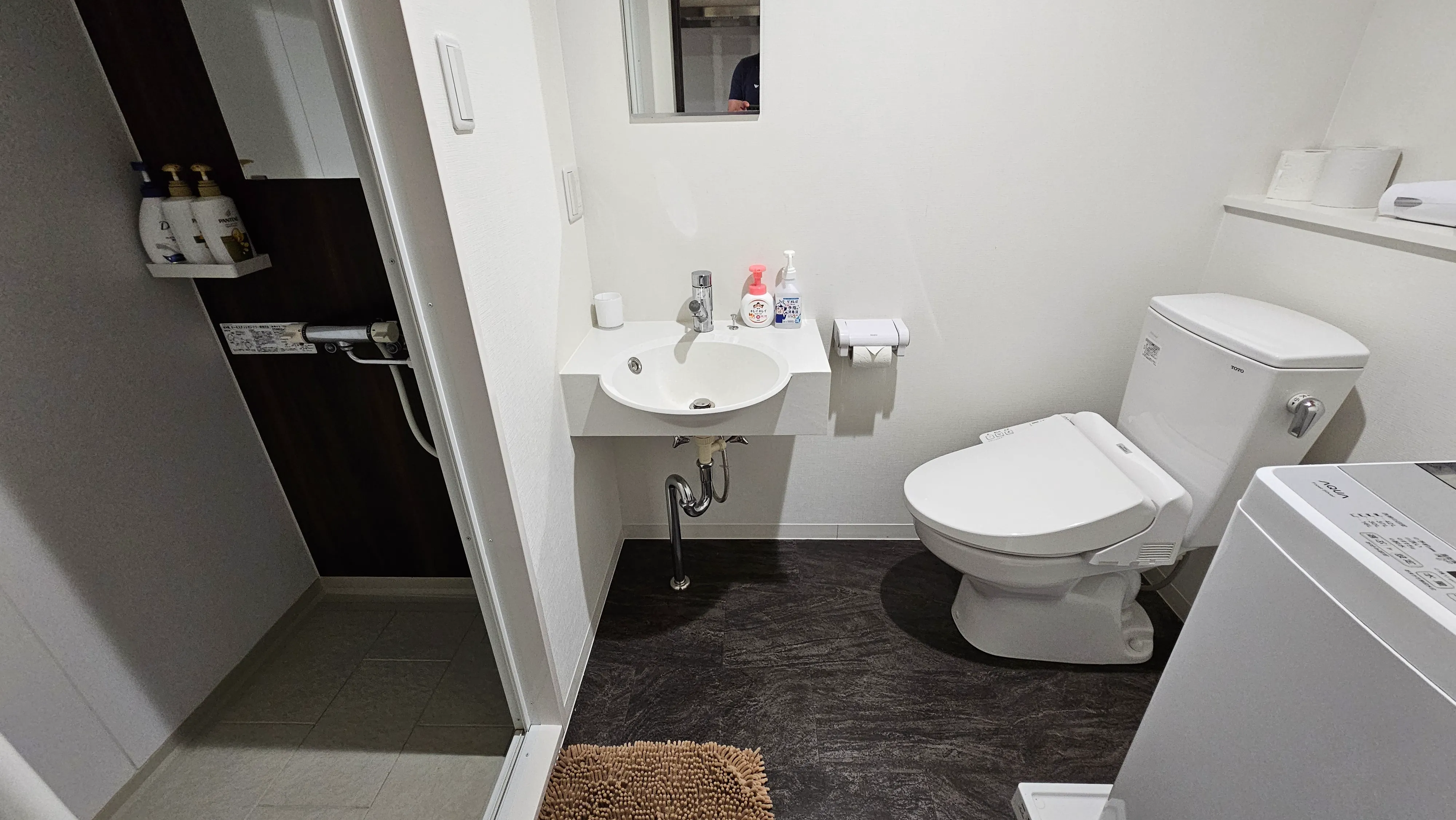 A small, modern bathroom with a white toilet, a sink with toiletries, a wall mirror, and a brown mat on the dark floor. An open doorway reveals a shower area with bottles on a shelf.