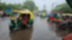 Rain-soaked street with colorful auto rickshaws and a motorcyclist. Pedestrians and blurred buildings in the background. Text reads "BLOW HORN."