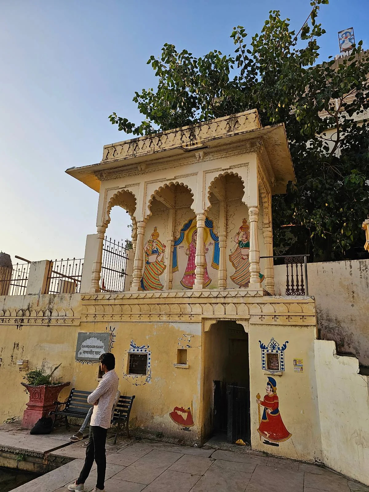 A person stands in front of a yellow building with ornate arches and colorful painted figures on the wall, likely depicting traditional Indian attire, with trees and a clear sky in the background.