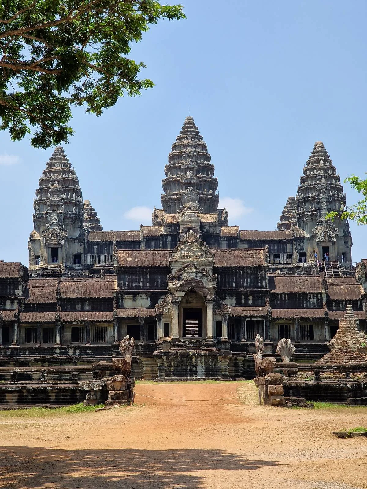 Angkor Wat temple in Cambodia, featuring intricate stone architecture, multiple towers, and steps leading up to the main entrance, with a blue sky and tree branches partly visible above.