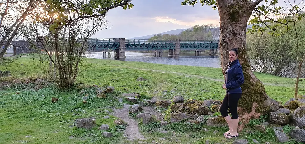 Woman in a navy jacket stands by a tree in a riverside park at sunset, with a green bridge and calm water behind her.