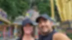 Couple in black caps smiling in front of colorful temple stairs and golden statue, surrounded by lush greenery, creating a joyful scene.