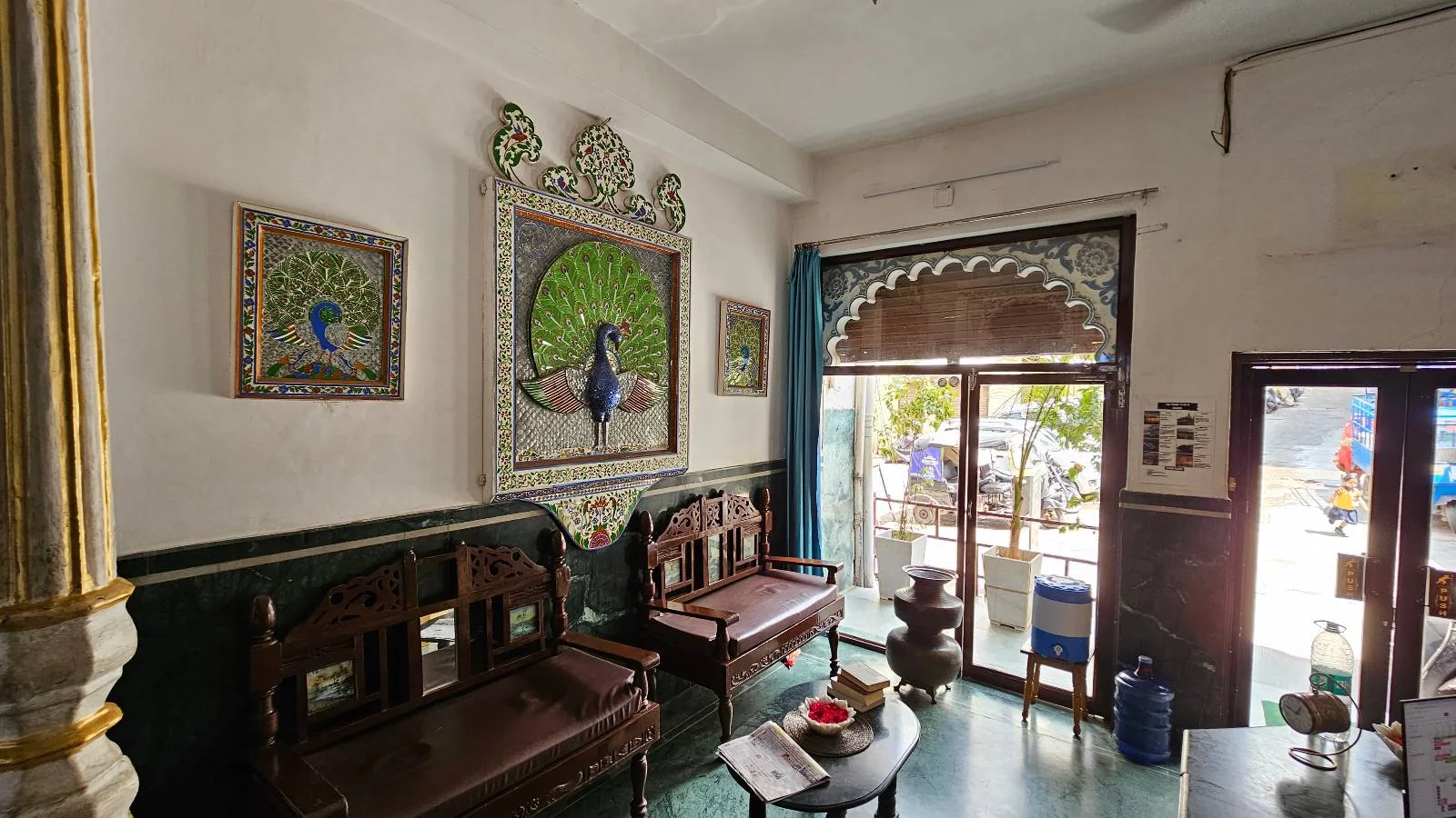 A cozy room with wooden benches, peacock-themed wall art, and a large arched window letting in daylight. There is a round table with cups, water dispensers, and decorative details on the walls and ceiling.