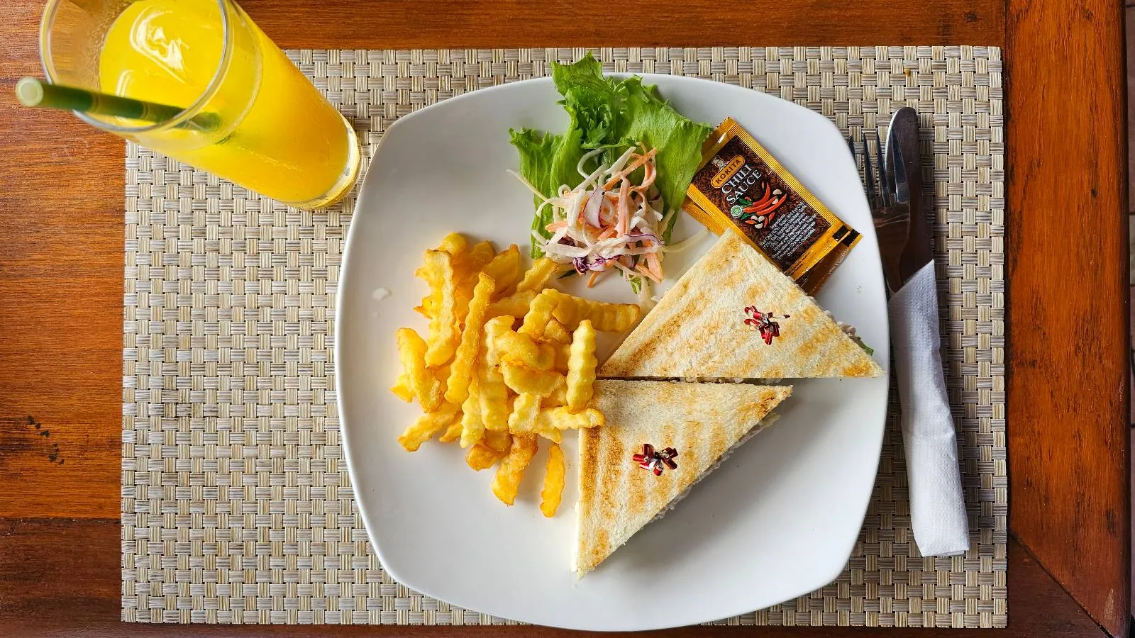 A plate with two grilled sandwiches, crinkle-cut fries, and a small salad garnish. A glass of orange juice with a straw and a knife and fork with a napkin are on the side. The setting is on a wooden table with a woven placemat.