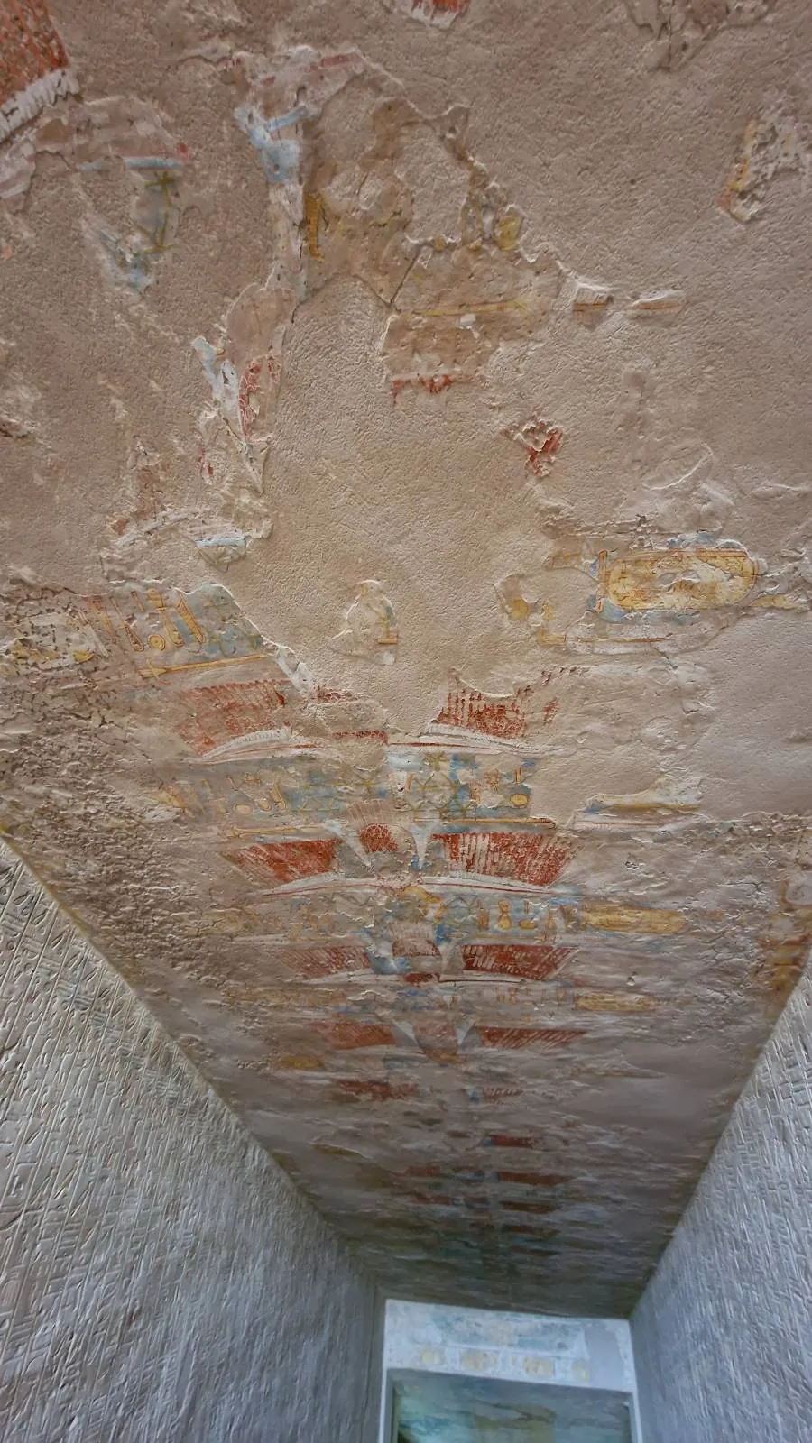 A narrow passageway with stone walls leads to a small rectangular entrance. The ceiling features worn, faded frescoes with hints of red, yellow, and blue, giving the area an ancient, historical appearance. Light filters in from the opening.