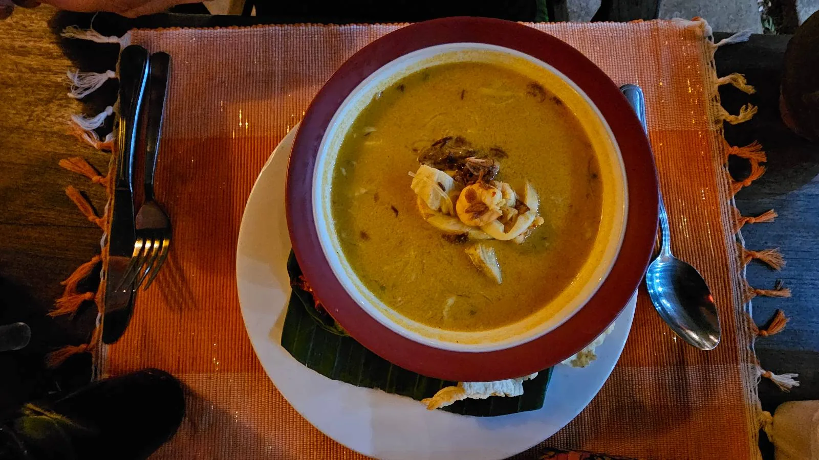 A bowl of yellow soup garnished with a dollop of cream is placed on a red-bordered plate. It sits on a table with a woven placemat. A fork and knife are on the left, and a spoon on the right.