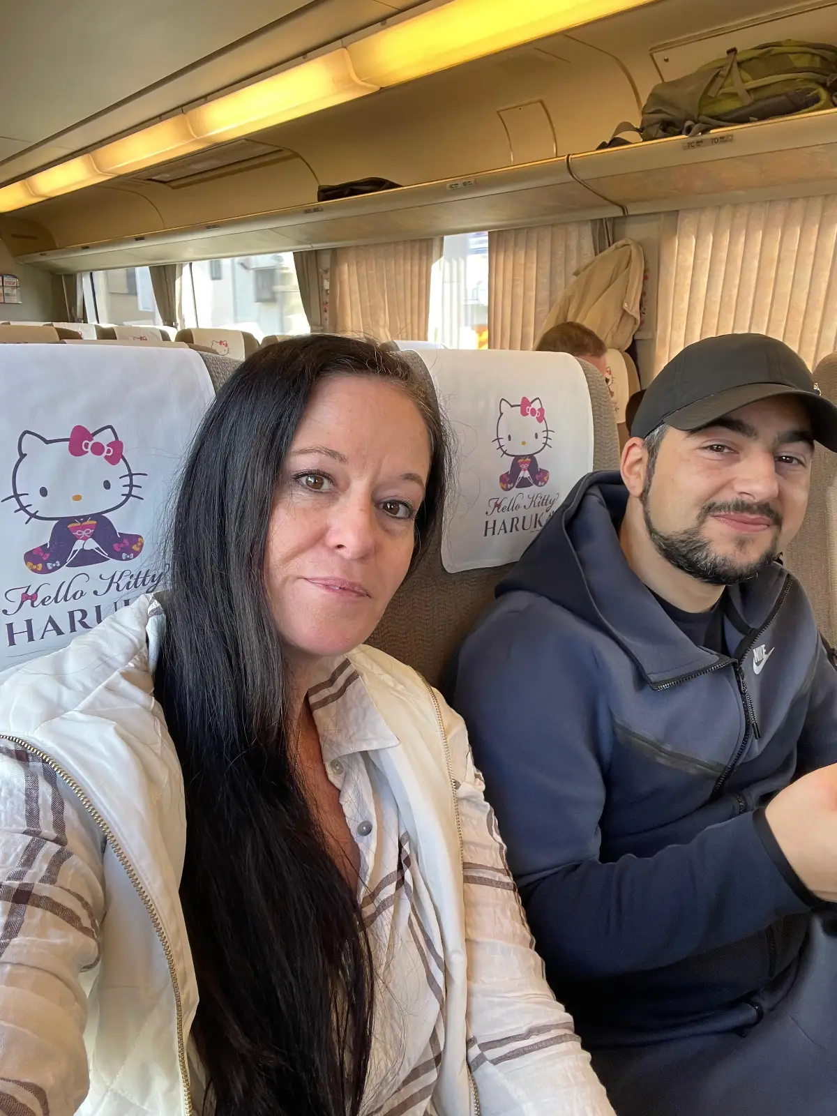 Two people sit side by side on a train with Hello Kitty-themed seat covers. The woman in front has long dark hair and wears a light jacket, while the man beside her wears a dark cap and smiles at the camera.