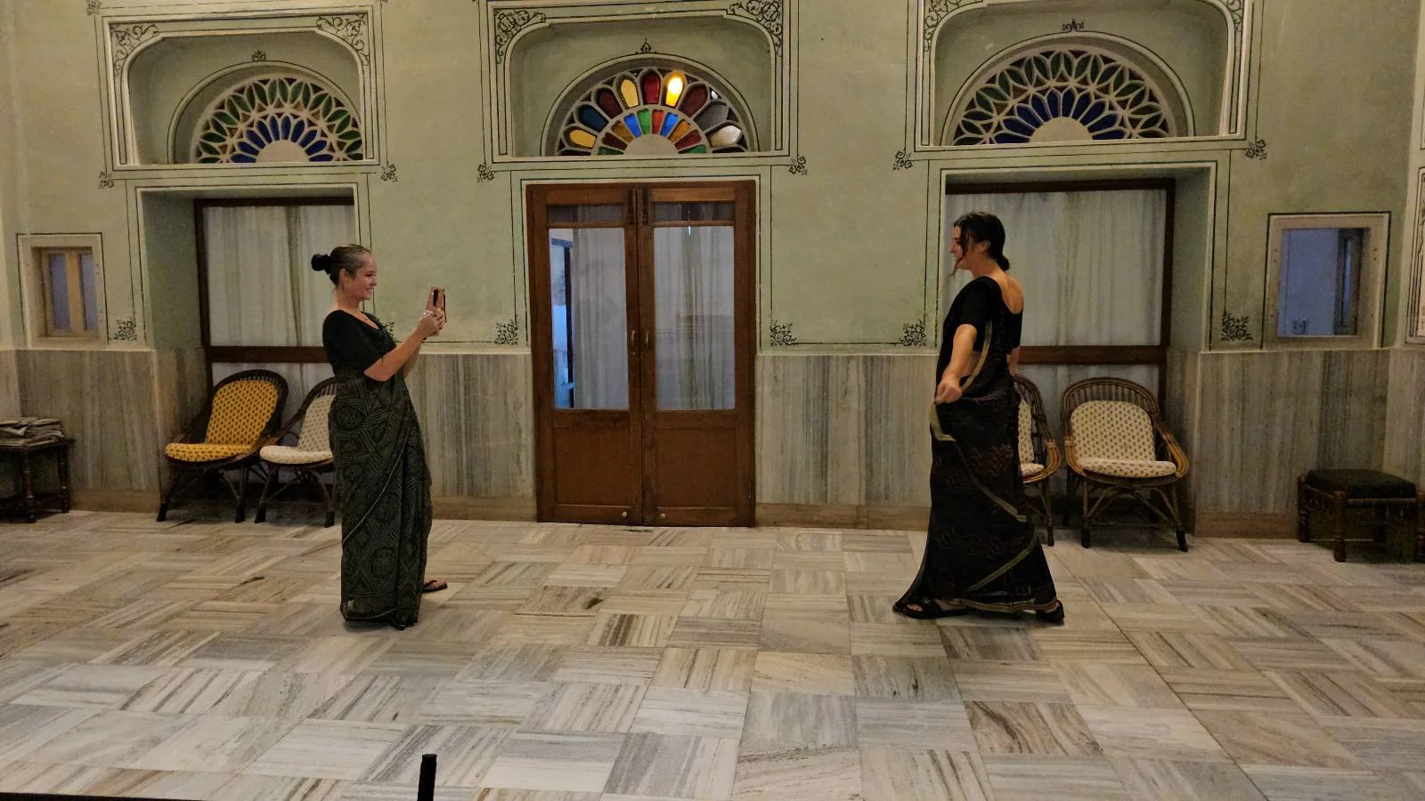 Two women in traditional clothing face each other in a spacious, ornate room with marble floors and arched windows. One woman is taking a photo of the other with her phone.