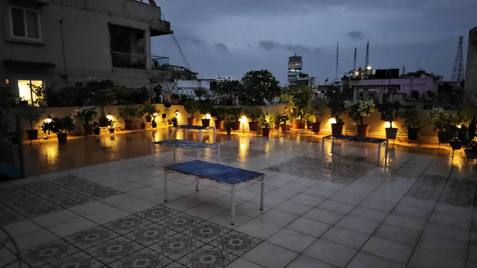 A rooftop patio with a white table and blue chairs overlooks a cityscape at dusk. Tall buildings and city lights are visible in the background under a cloudy evening sky.