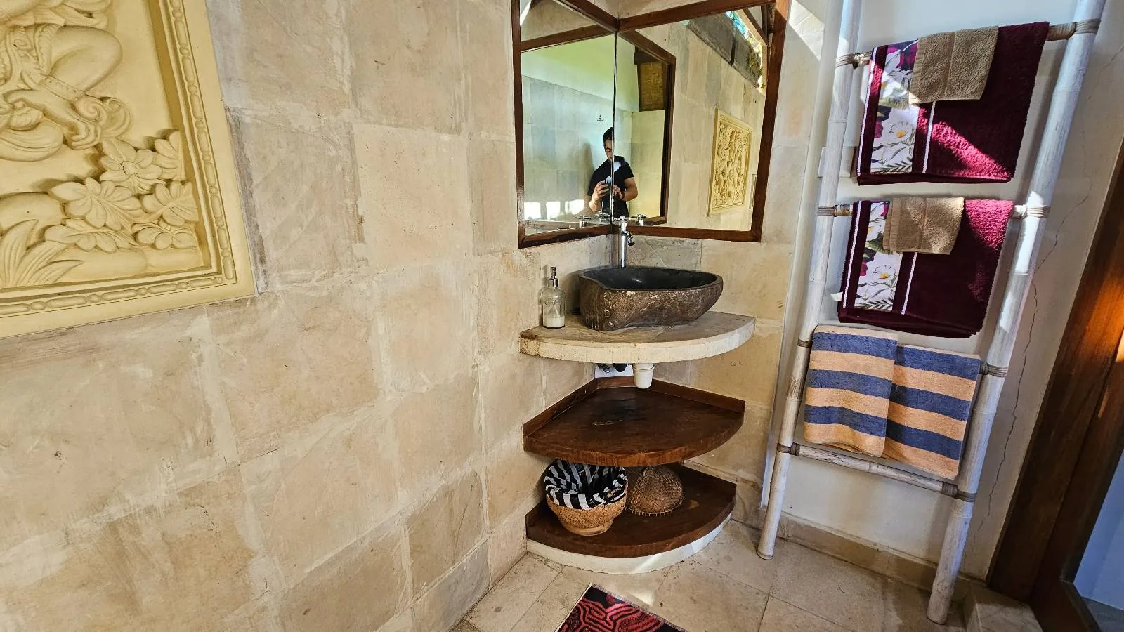 A small bathroom with stone walls, a corner sink with a dark stone basin, shelves underneath holding baskets, a towel rack with folded towels, and a mirror reflecting part of the room.