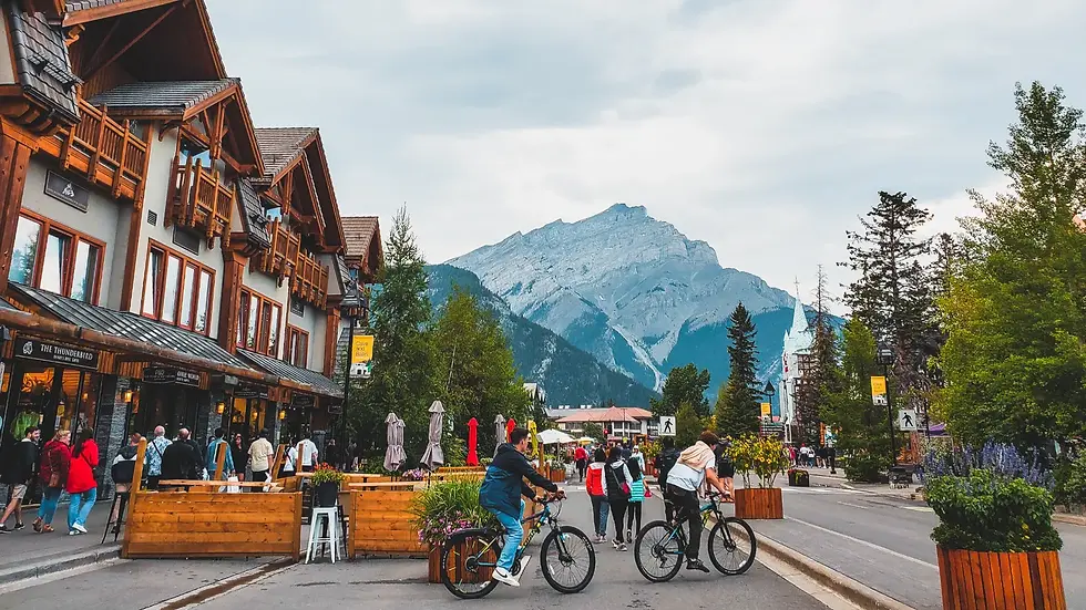 Street scene with people walking and cycling in a town surrounded by mountains, featuring wooden buildings and planters.