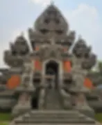 Ornate stone temple with intricate carvings, flanked by two statues on platforms. Central staircase leads to entrance. Overcast sky above.