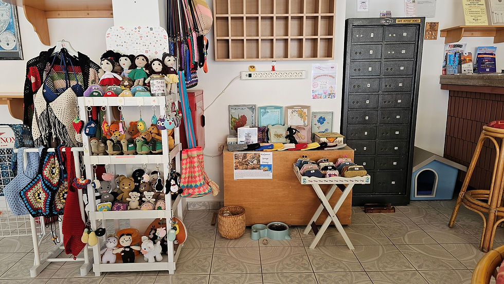 Crochet dolls and keychains on a white shelf in a cozy shop with a black safe deposit box and colorful textiles. Patterns and crafts visible.